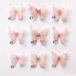 May include: A collection of nine individual butterfly stickers arranged in a grid, each featuring soft watercolour-style pink and peach tones. Each butterfly is labelled with a unique black number from 42 to 50, showcasing distinct wing patterns.