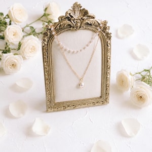 May include: A gold-toned layered necklace featuring a delicate chain with small white beads and a second chain with a single central pearl pendant. The necklace is displayed on a cream-coloured fabric insert within an ornate, antique-style gold frame.