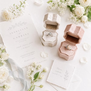 May include: A wedding invitation suite for Isabelle et Gabriel with a cream-colored paper card, two velvet ring boxes in beige and dusty rose, each containing a ring, surrounded by white roses and lilac sprigs on a white surface.