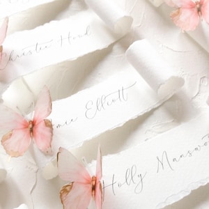 May include: A collection of white paper scrolls with deckled edges, each featuring handwritten names in black ink. The scrolls are decorated with delicate, light pink butterflies with gold-toned wing edges, arranged in a repeating pattern.