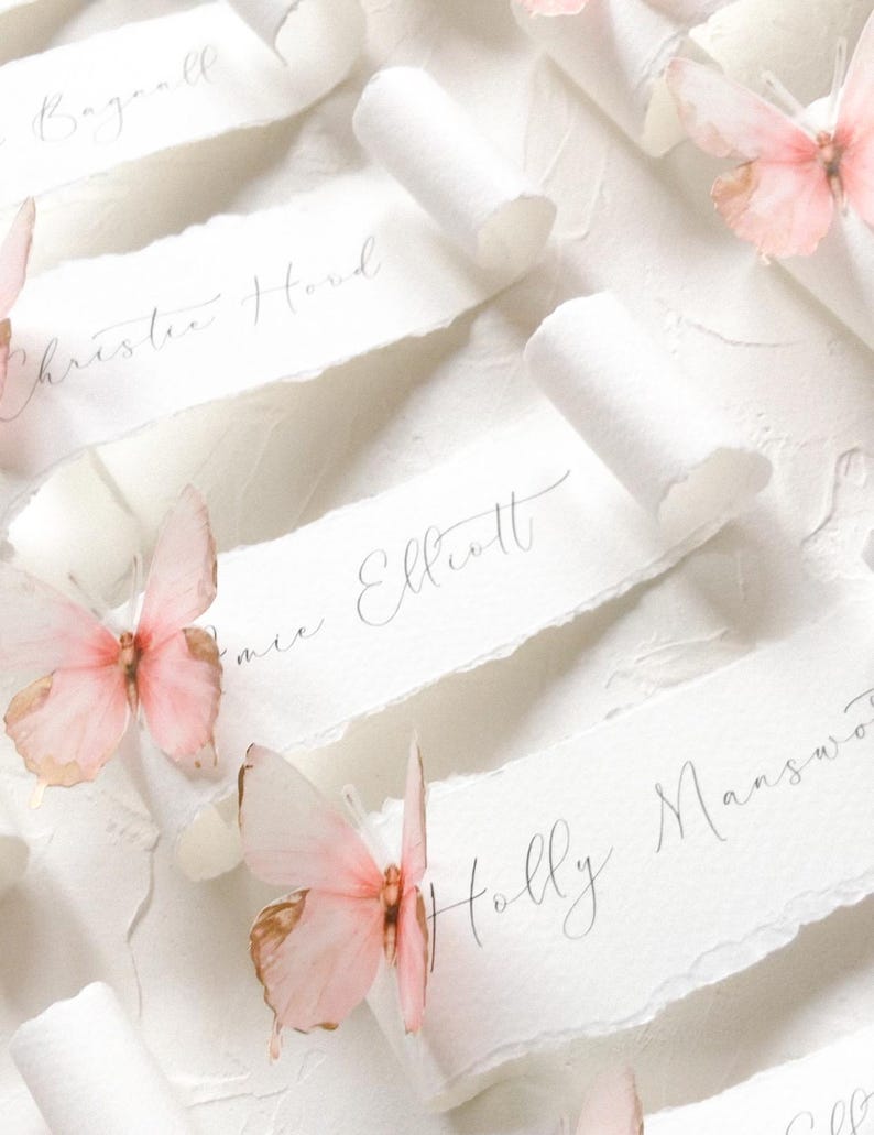 May include: A collection of white paper scrolls with deckled edges, each featuring handwritten names in black ink. The scrolls are decorated with delicate, light pink butterflies with gold-toned wing edges, arranged in a repeating pattern.