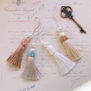 May include: Four decorative tassels in shades of beige, light grey, and white, each featuring a single white pearl bead and a textured knot detail at the top. A copper-coloured metal skeleton key is positioned to the right of the tassels.