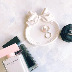 Pode incluir: Uma bandeja de joias branca em forma de laço contém dois anéis, um com uma pedra rosa. Uma caixa de anéis de veludo preto exibe vários anéis. Também estão presentes dois frascos de perfume com detalhes em rosa e preto, com o texto "narciso rodriguez for her".