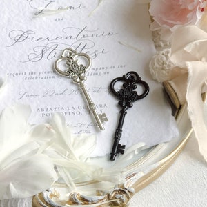 K&ouml;nnte beinhalten: Zwei verzierte Skelettschl&uuml;ssel aus Silber und Schwarz mit Kronendetails, die auf einem wei&szlig;en Papier mit Hochzeitseinladungstext liegen. Der Text lautet "Artemisia Lomi und Pierantonio Stitesi bitten um die Ehre Ihrer Gesellschaft bei der Feier ihrer Hochzeit am 22. Juni 202 in Abbazia La Celiara, Portofino, Italien."