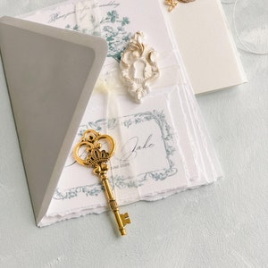 K&ouml;nnte beinhalten: Ein grauer Umschlag mit einem goldenen Schl&uuml;ssel und einem wei&szlig;en Dekorationsst&uuml;ck mit einem Schl&uuml;sselloch. Der Umschlag liegt auf einem Stapel wei&szlig;en Papiers mit blauen Blumenmustern und dem Text "Jake Juni 2023".