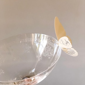 Puede incluir: Una copa de cristal transparente con un patr&oacute;n floral grabado en el cuenco. Una mariposa de papel de color crema est&aacute; sujeta al borde de la copa. La mariposa presenta el nombre Emily en letra cursiva decorativa, impreso en tinta negra.