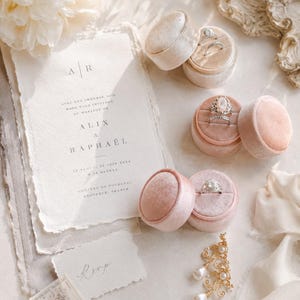 Könnte beinhalten: Ein Hochzeitseinladungsset mit handgeschöpftem Papier mit dem Text A|R Alix & Raphael, drei samtige Ringkästchen in Rosa- und Beigetönen mit Diamantringen, eine kleine RSVP-Karte, eine Briefmarke und goldene Ohrringe mit Perlen.