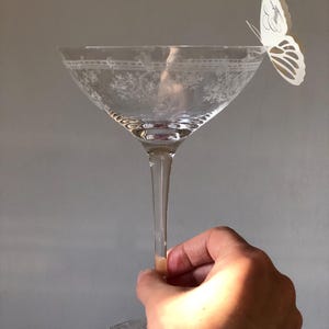 Puede incluir: Una copa de cristal transparente con un patr&oacute;n floral grabado alrededor del borde y una mariposa de papel blanco sujeta al borde. La mariposa tiene el nombre Emily escrito en una elegante letra cursiva negra sobre su ala.