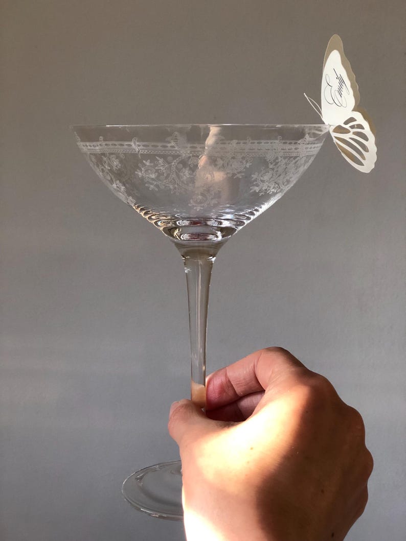 Puede incluir: Una copa de cristal transparente con un patr&oacute;n floral grabado alrededor del borde y una mariposa de papel blanco sujeta al borde. La mariposa tiene el nombre Emily escrito en una elegante letra cursiva negra sobre su ala.