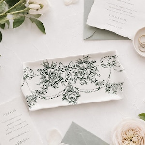 May include: A white ceramic tray with a dark green floral pattern of roses and ribbons. Wedding stationery and flowers are arranged around the tray. The tray is rectangular in shape.