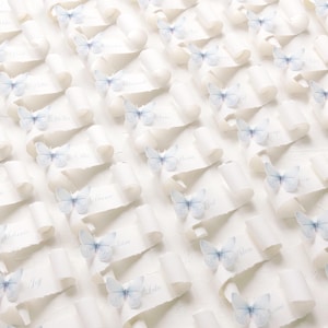 May include: A collection of white paper scrolls arranged in a grid pattern, each featuring a small, light blue butterfly accent. Each scroll displays a printed name in a cursive font, creating a uniform display of individual place cards.