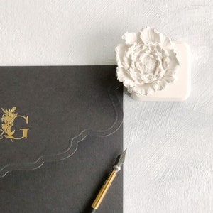 May include: A black envelope with a gold floral design and the letter "G" on the front. A white ceramic flower sits on a white surface next to the envelope. A gold and black calligraphy pen is on the envelope.