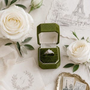 Peut inclure: Une bague argentée avec une pierre centrale en forme de poire et des pierres d'accent plus petites, présentée dans un écrin octogonal en velours vert olive. La boîte est ouverte, révélant une doublure blanche, près de roses blanches et papeterie.