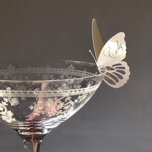 Puede incluir: Una copa de cristal transparente con un patr&oacute;n floral grabado alrededor del borde, con una tarjeta de sitio de papel blanco en forma de mariposa sujeta al borde. La mariposa tiene el nombre Emily impreso en fuente cursiva negra en el ala.