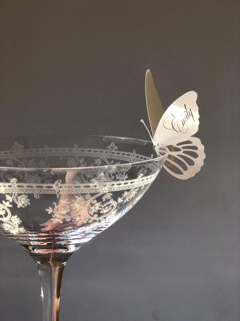 Puede incluir: Una copa de cristal transparente con un patr&oacute;n floral grabado alrededor del borde, con una tarjeta de sitio de papel blanco en forma de mariposa sujeta al borde. La mariposa tiene el nombre Emily impreso en fuente cursiva negra en el ala.