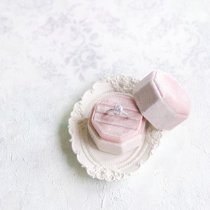 May include: A silver ring with a central oval-cut stone and small accent stones on the band, resting inside an octagonal light pink velvet ring box. The box sits on a small, ornate, cream-colored oval tray with a decorative floral-patterned edge.