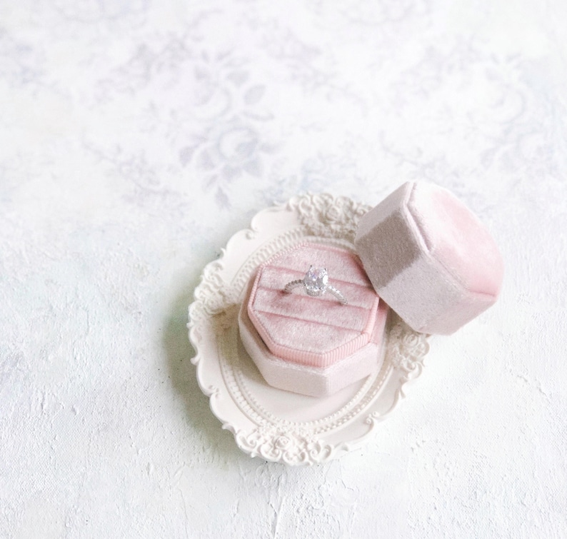 May include: A silver ring with a central oval-cut stone and small accent stones on the band, resting inside an octagonal light pink velvet ring box. The box sits on a small, ornate, cream-colored oval tray with a decorative floral-patterned edge.