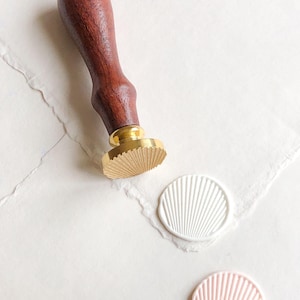 Puede incluir: Un sello de cera con mango de madera y cabeza de sello dorada, junto a dos sellos de cera blancos y rosas con diseño de abanico. El sello está sobre una superficie de papel blanco.