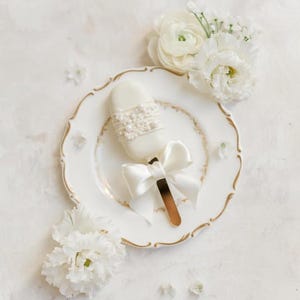 Op de afbeelding: Een witte chocolade-ijsje op een stokje met een wit lint strikje en parel accenten op een wit bord met een gouden rand. Het bord is omringd door witte bloemen.