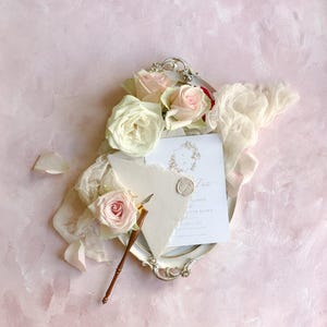May include: An elegant wedding invitation suite featuring a white card with gold script and a wax seal, nestled on a silver tray with roses and a vintage pen. Soft pink and white roses add a romantic touch.