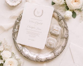 May include: A wedding invitation with the names Victoria Elizabeth and Christopher James, displayed on a silver platter with two ring boxes. The invitation has a wreath design and is surrounded by white roses and a white ribbon.
