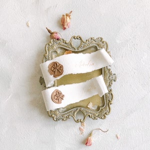 Może przedstawiać: Dwa białe zwoje papieru z pieczęciami woskowymi w ozdobnej złotej ramie. Zwoje są opisane nazwami "Aurelia" i "Justin".