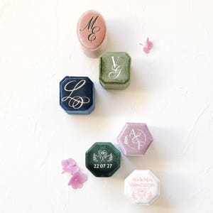 May include: A collection of six velvet ring boxes in various shapes and colours, including dusty rose, olive green, navy blue, mauve, dark green, and white. Each box features custom white or gold monogram lettering, dates, or swan illustrations.