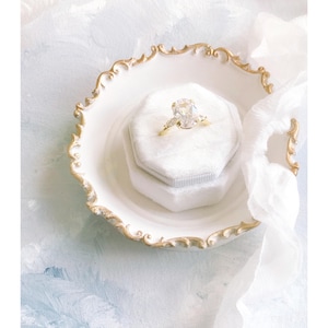 Puede incluir: Un anillo de compromiso de oro con un gran diamante de talla ovalada y piedras laterales más pequeñas descansa en una caja de terciopelo blanco. La caja está dentro de un plato blanco adornado con ribete dorado. Una tela blanca suave se extiende alrededor del plato, sobre un fondo pintado de azul claro y blanco.