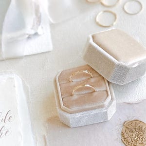 May include: Two thin gold rings are displayed inside a beige velvet hexagonal ring box with its lid resting beside it. Several additional thin gold rings are scattered nearby, alongside a piece of stationery with the text, With great pleasure YOU ARE INVITED TO CELEBRATE, and a gold wax seal.