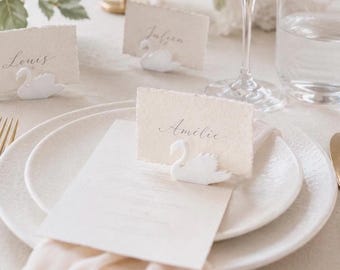 May include: A white ceramic swan place card holder featuring a slot for a rectangular paper card with deckled edges. The card displays the name Amélie in elegant cursive script. The swan is positioned on a white plate with a light fabric napkin.
