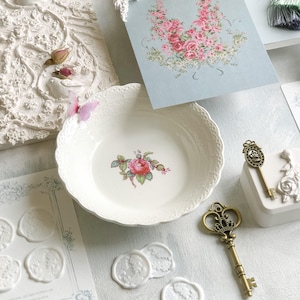 K&ouml;nnte beinhalten: Wei&szlig;e Keramikschale mit einem Blumendesign mit einer rosa Rose und gr&uuml;nen Bl&auml;ttern. Die Schale hat einen gewellten Rand und ist umgeben von wei&szlig;en Wachssiegeln, einem goldenen Schl&uuml;ssel und einer Karte mit einem Blumendesign.
