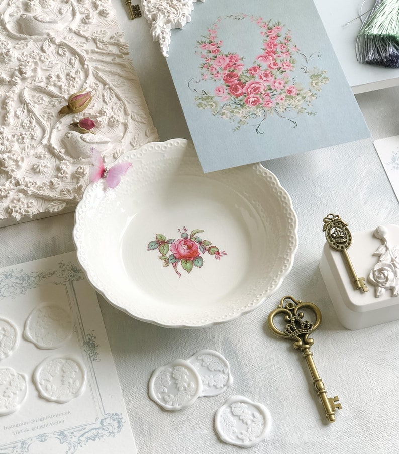K&ouml;nnte beinhalten: Wei&szlig;e Keramikschale mit einem Blumendesign mit einer rosa Rose und gr&uuml;nen Bl&auml;ttern. Die Schale hat einen gewellten Rand und ist umgeben von wei&szlig;en Wachssiegeln, einem goldenen Schl&uuml;ssel und einer Karte mit einem Blumendesign.