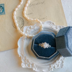 Pode incluir: Um anel de prata com uma grande gema transparente cercada por pedras menores, apresentado em uma caixa de anel de veludo azul. A caixa está colocada em um prato decorativo branco e adornado. Um colar de pérolas está drapeado sobre um envelope vintage.