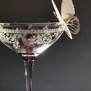 Puede incluir: Una copa de cristal transparente con un intrincado patr&oacute;n floral blanco grabado alrededor del borde. Una delicada mariposa de papel de color blanco roto est&aacute; posada en el borde de la copa, proyectando una suave sombra sobre la superficie.