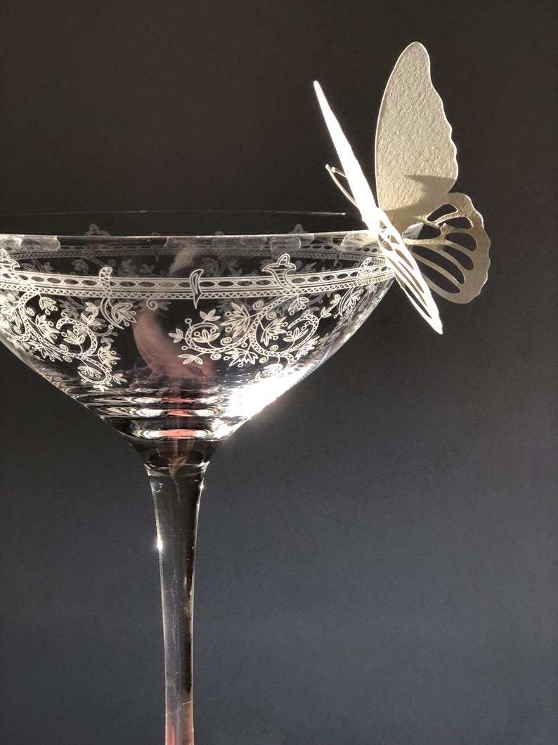 Puede incluir: Una copa de cristal transparente con un intrincado patr&oacute;n floral blanco grabado alrededor del borde. Una delicada mariposa de papel de color blanco roto est&aacute; posada en el borde de la copa, proyectando una suave sombra sobre la superficie.