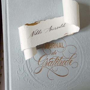 May include: A light blue journal with gold embossed text that reads "JOURNAL de Gratitude" and a decorative border. Resting on the cover is a piece of white, deckle-edged paper with the name "Nikki Amirsaleh" written in black calligraphy.