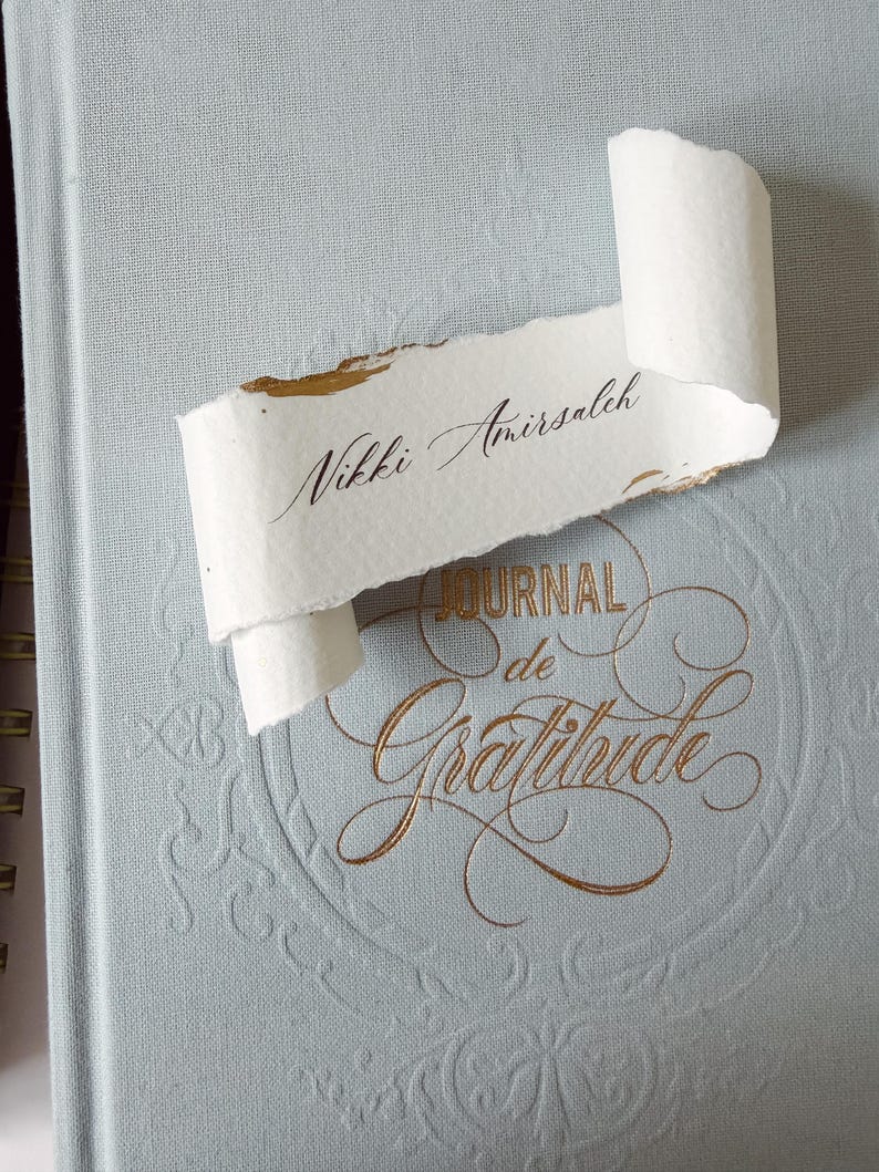May include: A light blue journal with gold embossed text that reads "JOURNAL de Gratitude" and a decorative border. Resting on the cover is a piece of white, deckle-edged paper with the name "Nikki Amirsaleh" written in black calligraphy.