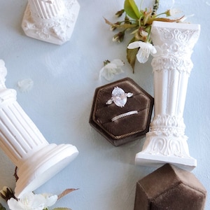 May include: A white oval-shaped engagement ring with a diamond center stone and smaller diamonds on the band. The ring is in a brown velvet ring box with a white satin lining. The ring box is on a white surface.