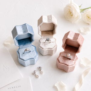 以下が含まれることがあります： ライトブルー、ベージュ、ダスティピンクのベルベット製リングボックス3つ。それぞれにシルバーリングが収められています。白いパールの付いたシルバーのドロップイヤリング2つが近くに置かれています。AURÉLIE et DAMIEN 22.06.2025と書かれた白いカードが隅に見えます。