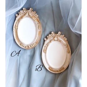 Puede incluir: Dos marcos de fotos ovalados y ornamentados, con acabado dorado y blanco. Los marcos tienen detalles decorativos y están etiquetados como "A" y "B". Se muestran sobre una tela suave azul-gris.