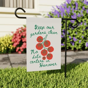 May include: A garden flag with the text "Keep our gardens clean" and "No data centers in Hanover" in green script. A cluster of red tomatoes is in the center. The flag is displayed on a black metal stand in a garden.