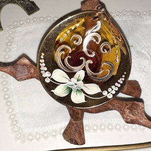 Puede incluir: Un pequeño cuenco de vidrio color ámbar con diseños florales pintados en oro y blanco se asienta sobre un soporte de madera con tres patas. El cuenco tiene un centro de vidrio rojo.