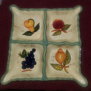 May include: A square, off-white ceramic serving dish with four sections, each decorated with a different fruit: a pear, an apple, grapes, and a citrus fruit. The dish has a light green border and a decorative edge.