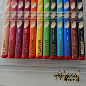 Könnte beinhalten: Ein Set mit 12 Buntstiften in einem durchsichtigen Plastiketui. Die Stifte sind mit dem Namen "HAMZA" und einer Nummer beschriftet. Die Stifte sind in einer Reihe angeordnet, wobei der rote Stift oben und der braune Stift unten ist.