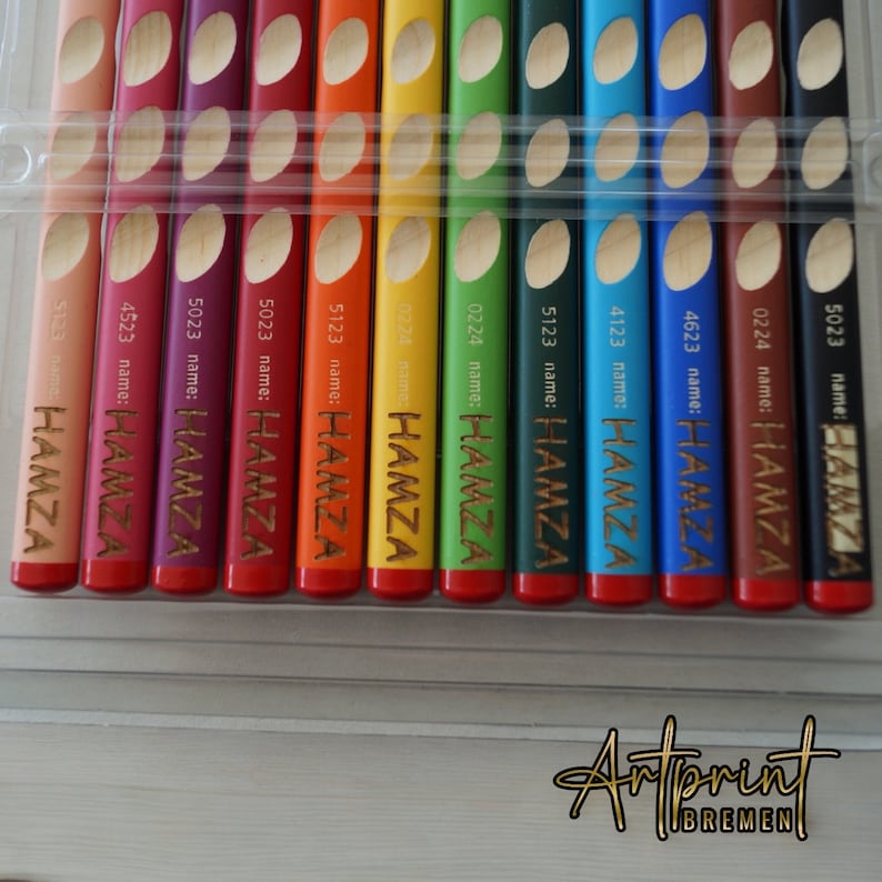 K&ouml;nnte beinhalten: Ein Set mit 12 Buntstiften in einem durchsichtigen Plastiketui. Die Stifte sind mit dem Namen "HAMZA" und einer Nummer beschriftet. Die Stifte sind in einer Reihe angeordnet, wobei der rote Stift oben und der braune Stift unten ist.