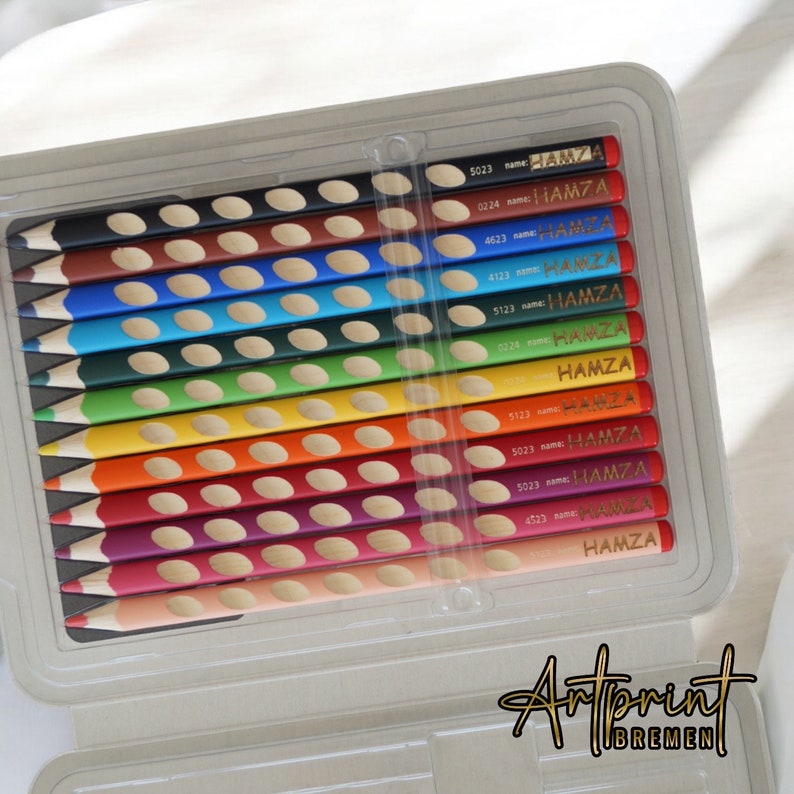 K&ouml;nnte beinhalten: Ein Set mit 12 Buntstiften in einem durchsichtigen Plastiketui. Die Stifte sind in Regenbogenfarben angeordnet und jeder Stift hat ein goldenes Etikett mit dem Namen "HAMZA" darauf.