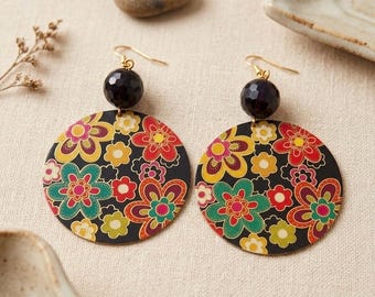 May include: A pair of dangle earrings featuring large, flat, circular discs with a black background and a colorful floral pattern in shades of red, yellow, green, and pink. Each earring is topped with a faceted, dark, spherical bead and a gold-tone hook.