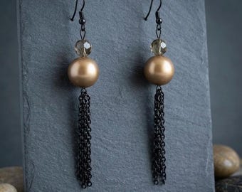 May include: A pair of dangle earrings featuring a black metal hook, a small faceted translucent bead, a large smooth matte gold sphere, and a multi-strand black chain tassel. The earrings are suspended from a dark gray textured display stand.