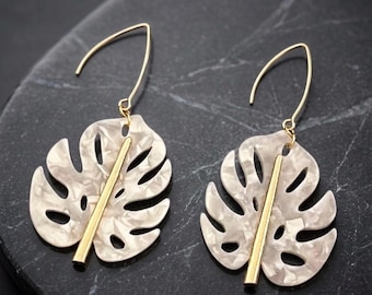 May include: A pair of dangle earrings featuring cream-colored, marbled monstera leaf pendants. Each leaf has a central gold-tone metal spine and is suspended from a gold-tone curved ear wire hook, creating a botanical design for the ears.