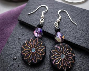 May include: A pair of dangle earrings featuring round black discs with an iridescent, star-shaped floral pattern in shades of purple and gold. Each earring includes a small black bead and a faceted purple crystal bead suspended from a silver hook.